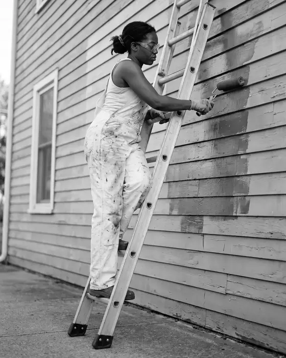 A painter on an extension ladder rolling fresh paint onto the upper siding